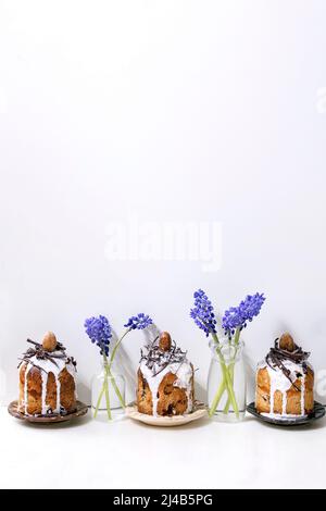 Homemade traditionla small Easter kulich cakes with chocolate nests and eggs on plates in row decorated with muscari flowers over white marble table. Stock Photo