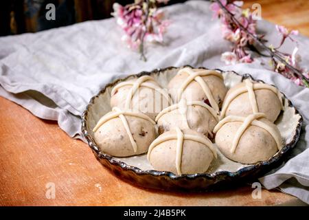 Raw unbaked buns. Ready to bake homemade Easter traditional hot cross buns in ceramic baking dish over orange stone table with linen cloth and pink fl Stock Photo