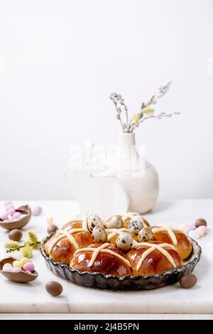 Homemade Easter traditional hot cross buns in ceramic dish with blossom flowers, chocolate candy and quail eggs on white marble table. Easter holiday Stock Photo