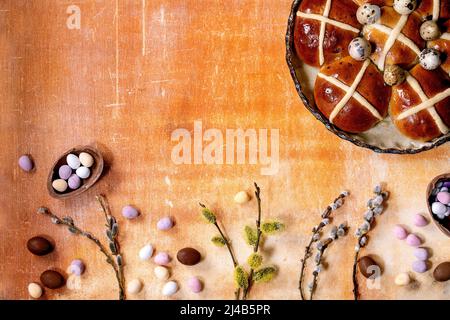 Homemade Easter traditional hot cross buns in ceramic dish with blossom willow branches and chocolate eggs candies over orange stone background. Flat Stock Photo