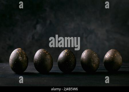 Black Easter concept. Bio colored black eggs with golden spots in row on dark wooden background. Copy space Stock Photo