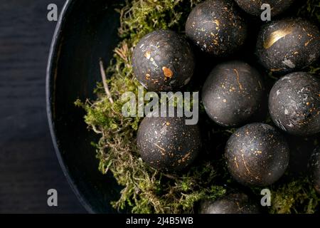 Black Easter concept. Bio colored black eggs with golden spots laying on wild moss in black ceramic bowl on dark wooden background. Top view Stock Photo