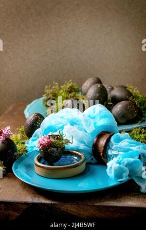 Easter table setting with empty turquoise ceramic plates, decorated by forest moss, black colored easter eggs and pink spring flowers in shell on wood Stock Photo