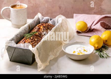 Homemade lemon cake with nuts and rosemary in baking dish, serving with fresh lemon, white sauce and coffee mug on white marble table. Stock Photo