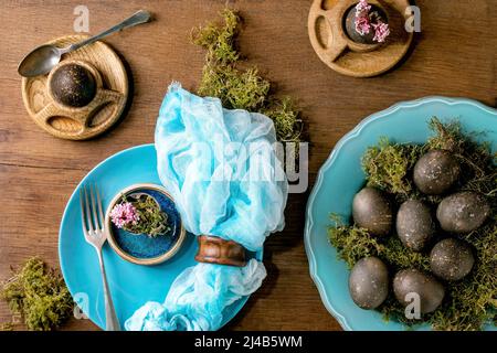 Easter table setting with empty turquoise ceramic plates and egg cups, decorated by forest moss, black colored easter eggs and pink spring flowers on Stock Photo
