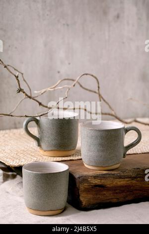 Set of empty grey texture craft ceramic coffee cups with dry branch on linen tablecloth with wooden board. Grey wall, copy space Stock Photo