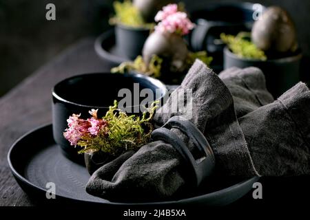 Easter table setting with empty black ceramic plates and mugs, decorated by forest moss, black colored easter eggs and pink spring flowers on wooden t Stock Photo