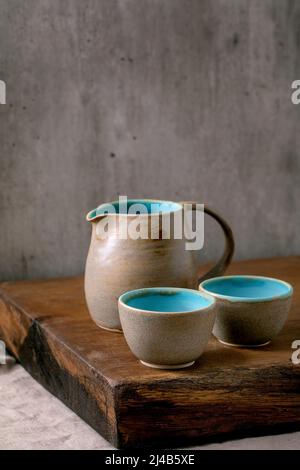 Set of empty turquoise grey textured craft ceramic jug and cups standing on wooden table with grey wall at background Stock Photo