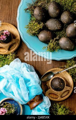Easter table setting with empty turquoise ceramic plates and egg cups, decorated by forest moss, black colored easter eggs and pink spring flowers on Stock Photo