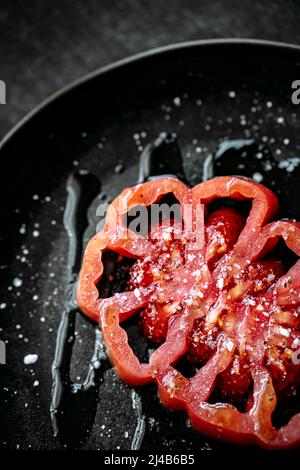 Close up of a slice of beef heart tomato, placed on a black plate, salt, pepper and olive oil, on a black textured background. view from above Stock Photo