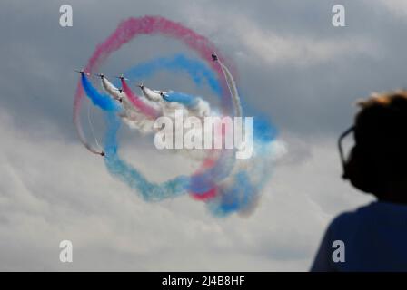 The RAF Red Arrows aerobatic team giving a display with two aircraft performing a roll around the remaining seven, all trailing colored smoke Stock Photo