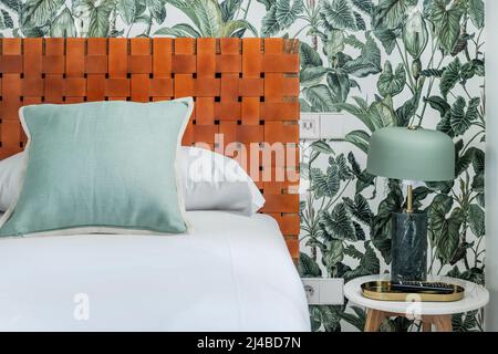 Detail image of a bedroom with woven brown leather headboard, sky blue cushions, wall with decorative wallpaper and green lampshade lamp Stock Photo