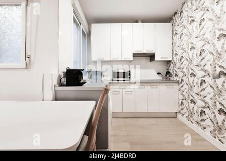 White kitchen with wooden furniture, small appliances on the counter, a wall with decorative paper and a white dining table with beech wood chairs Stock Photo
