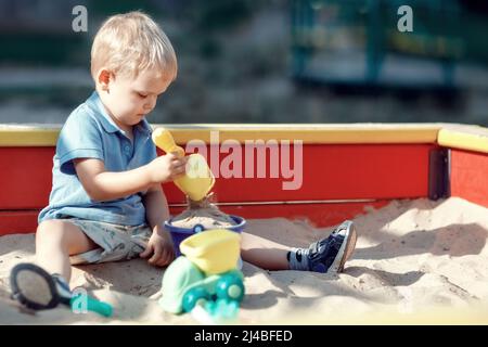 Toddler boy is playing in a red and yellow edge sandbox with sand toys. Accumulated baby with spade in hand is scooping sand into a blue bucket. Child Stock Photo