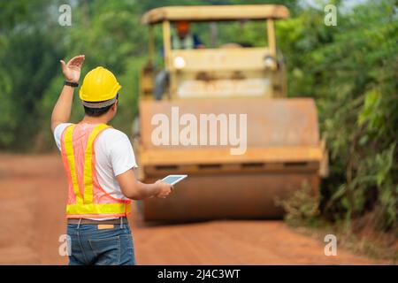 Asian civil engineer operate with tablet to control working soil compaction machine compacting soil under the road construction in the countryside. Stock Photo