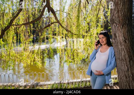 A smiling pregnant woman with makeup talks on her mobile phone and looks right at the camera while she holds her big pregnant stomach with her left hand. Stock Photo