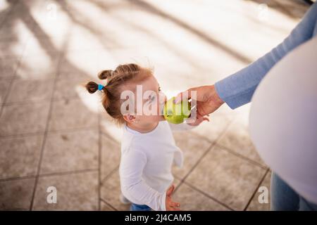 A cute caucasian little girl with ponytails stands and eats a green apple while a pregnant unrecognizable woman feeds her. Stock Photo