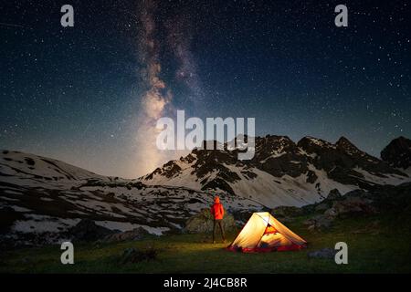 Hiking tourist standing near illuminated tent under milky way stars Stock Photo