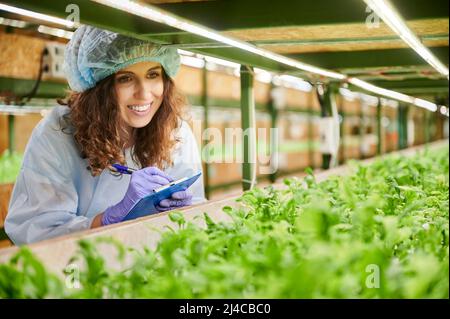 Female researcher in disposable cap looking at green leafy plants and smiling while writing on clipboard. Joyful woman in garden gloves checking plant growth and taking notes in greenhouse. Stock Photo