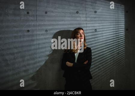 A dreamy senior brown-haired formal clothed caucasian woman stands in front of gray background in darkened room with crossed arms. Blins shadows. Stock Photo