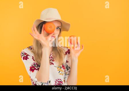 Youthful caucasian blonde girl in her 20s in a hat posing with two oranges or mandarins. Medium studio shot over orange background. High quality photo Stock Photo