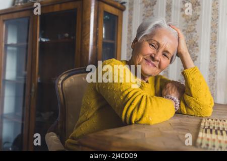 Happy glad about her long life gray-haired lady looking at camera and smiling while relaxing by wooden table and touching her hair. High quality photo Stock Photo