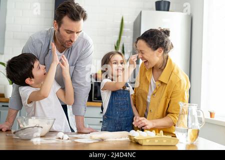 Daughter smearing nose of mom with flour and laughing. Happy family of four cooking in the kitchen together, parents and two kids making dough together, preparing pie or cookie, have a fun on weekend Stock Photo