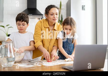 Middle-aged mother and her kids cooking together, kneads the dough, using rolling pin. Mom and kids spends leisure at home, baking cookies, looking tutorial at the laptop, enjoying weekend activity Stock Photo