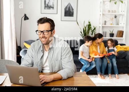 Happy father wearing glasses is working on the distance. Man using laptop for freelance work sitting at the table at home while family sitting peacefully together on the sofa Stock Photo