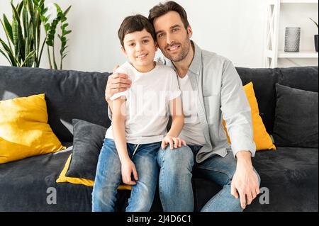 Portrait of cheerful father and son sitting on the sofa in cozy living room, looking at the camera and laughing, enjoying weekend at home. Happy together concept Stock Photo