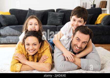 Happy multiracial family of four laying at the floor in cozy living room, parents and kids laughing while embracing with each other. Family feeling happy during the weekend Stock Photo