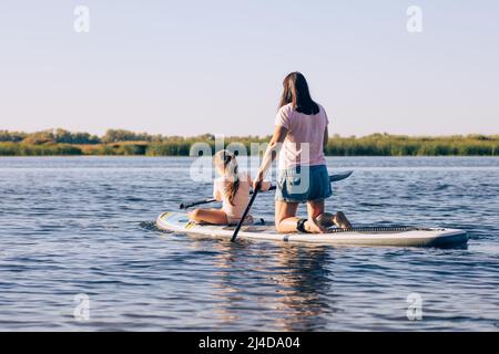 Mother and little daughter on sup board rowing together effortfully, exercising and having fun on lake with green reeds in background. Active Stock Photo