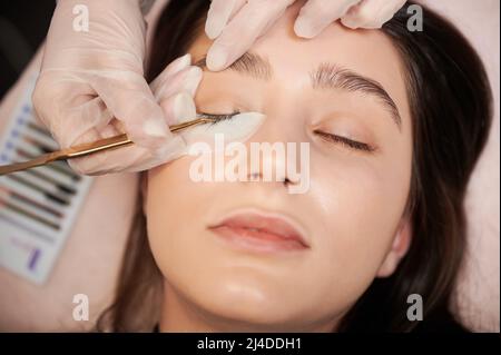 Close up of lash specialist in sterile gloves applying lash extensions with tweezers. Young woman with eye patch under lower eyelid keeping eyes closed while having eyelash extension procedure. Stock Photo