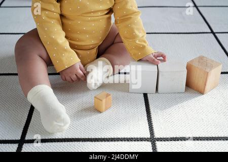 HIgh angle shot of of unrecognizable baby wearing yellow long-sleeve onesie and white socks sitting on floor touching wooden cubes Stock Photo