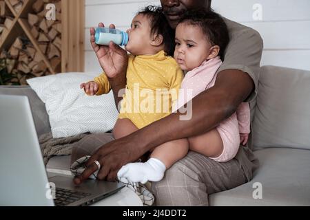 Modern African American father sitting on sofa in living room working on laptop and taking care of his baby daughters at the same time Stock Photo