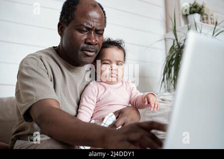 Portrait of modern mature African American man sitting on sofa with baby daughter on his lap working on laptop Stock Photo
