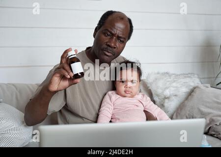 Modern mature Black man sitting on sofa in front of laptop with baby daughter on his lap having online consultation with pediatrician Stock Photo