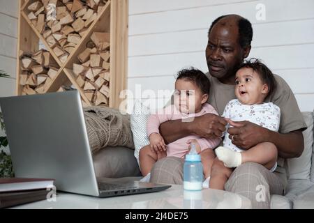 Portrait of modern mature African American father sitting on sofa in living room with twin babies on his lap watching movie in Internet on laptop Stock Photo