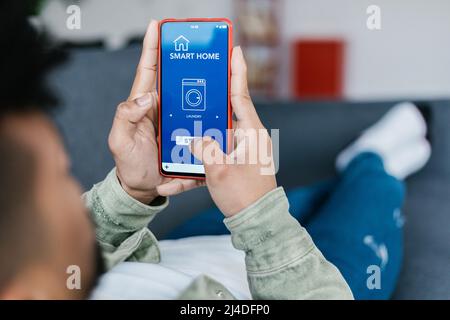 Young man lying on sofa and holding phone with app for controlling smart home Stock Photo