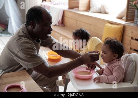 Modern mature African American man taking care of his baby daughters feeding them with fruit puree Stock Photo