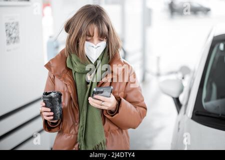 Woman in face mask holding smart phone and coffee cup while refueling car at gas station. Stock Photo
