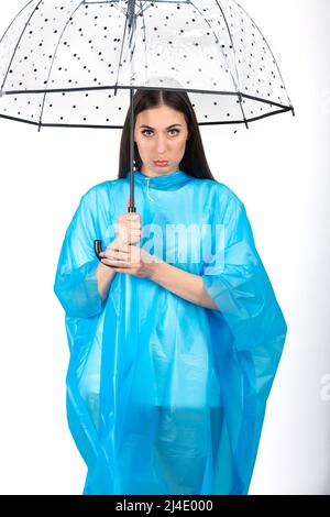 Studio shot of beautiful woman with long dark hairs wearing blue raincoat and hiding under transparent with black dots retro style umbrella in white b Stock Photo