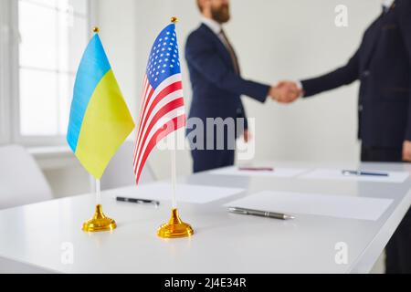 Flags of Ukraine and USA on table with two men in background exchanging handshakes Stock Photo