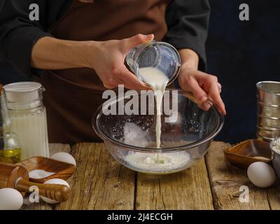 Cooking dough in a glass bowl by the hands of a professional chef on a wooden kitchen table against a dark background. The chef pours milk into a bowl Stock Photo