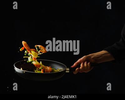 A professional chef prepares shrimp with vegetables and lemon slices in a frying pan on a black background. Levitation. Asian cuisine, healthy vegetar Stock Photo