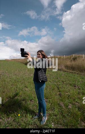 Beautiful hispanic woman blogger filming herself with her camera in her hand happy posing in the middle of the field on a cloudy afternoo Stock Photo