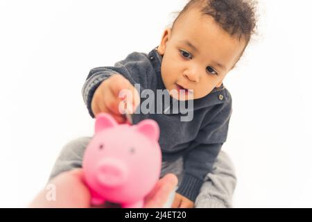 Adorable baby boy using pink piggy bank by putting coins there. Future and savings concept. Studio shot over white background. High quality photo Stock Photo