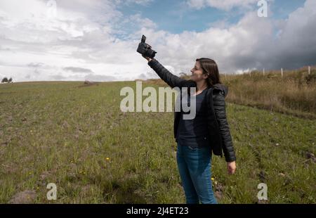 Beautiful hispanic woman blogger filming herself with her camera in her hand happy posing in the middle of the field on a cloudy afternoon Stock Photo