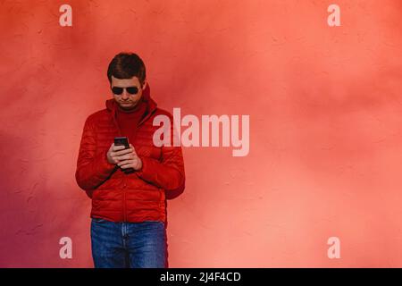 Young man in red sweater and jacket standing near the red wall, using his smart phone Stock Photo
