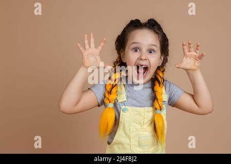 Portrait of indulging little girl having yellow kanekalon pigtails, frightening someone with hands and weird grimace wearing yellow jumpsuit and gray Stock Photo